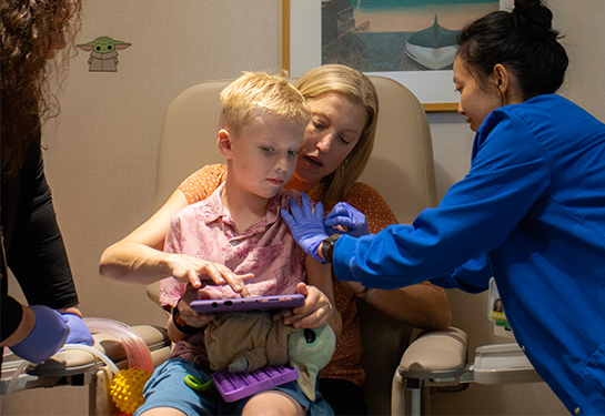 A mother with medium-length blonde hair holds her young son, also blonde, on her lap in a medical chair as a nurse and cleans the boy&#x2019;s arm.
