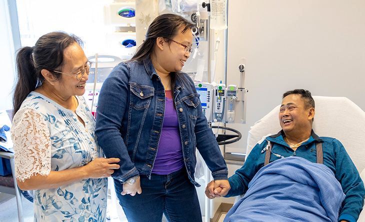 Asian American man resting in bed with IV in arm, holding hands with young Asian American female and older Asian American female.