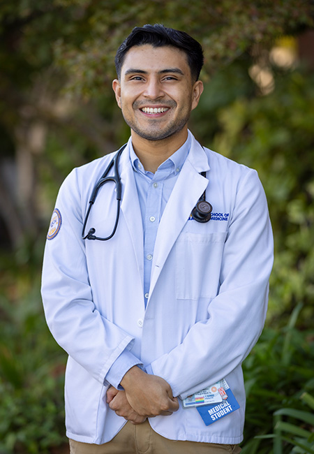 A young man with dark hair and white coat with the words “School of Medicine” on the lapel poses for the camera