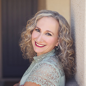 A woman with curly blonde hair, wearing a light green, short-sleeved blouse, leans against the pillar of a home, smiling. 