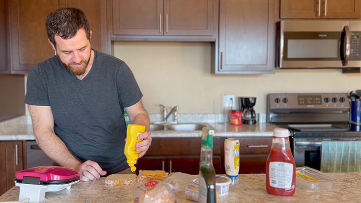 A man with dark hair and beard, in gray scrubs, assembles a sandwich on the kitchen island of an apartment 