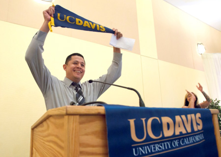 Godoy is holding a UC Davis banner in his two hands on his match day. Someone in the background is cheering and clapping.
