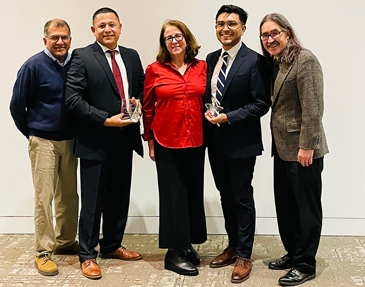 Five standing individuals smiling to the camera. Dr. Godoy (second from left) is holding his rectangular glass award. Andrés Maldonado (is second from right) is holding his star-shaped glass award. Interim Dean Susan Murin (in the center) is wearing a red short and a long black skirt.