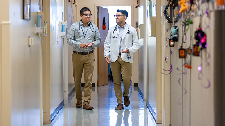 A man with short dark hair walks down a hallway, gesturing his hands with a younger man who is a medical student in white coat