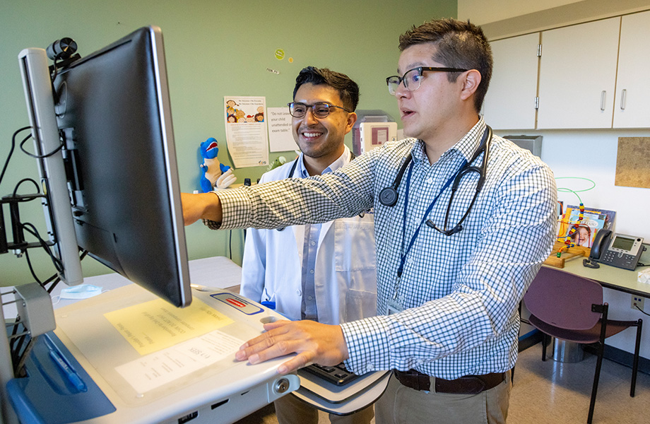 A doctor in a long-sleeve shirt points to a computer screen as his medical student mentee in white lab coat looks on and smiles