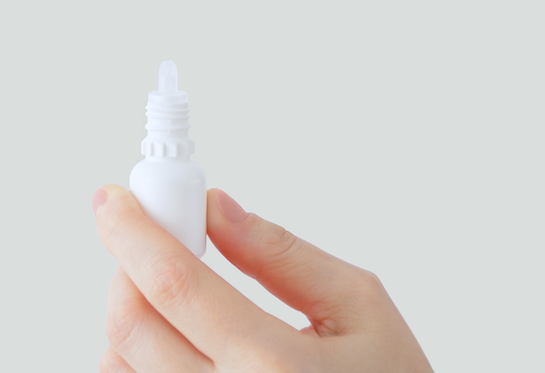 A hand holds a white eyedrop bottle against a light grey background. 