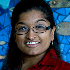 A woman with dark hair, wearing a maroon shirt and glasses, smiles for a portrait. 