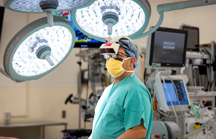 A surgeon wearing green scrubs, a cap and an augmented reality headset stands in an operating room beneath two giant overhead lights.