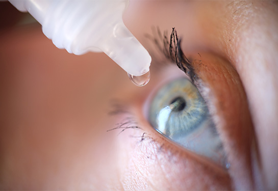 A transparent drop dangles from the tip of a white vial above a closeup of a human eye.