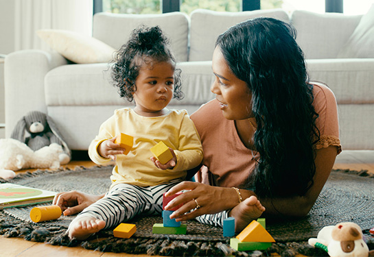 A woman with long dark hair wearing a peach-colored shirt lays on the floor next to an infant wearing black-and-white pants and a yellow shirt.