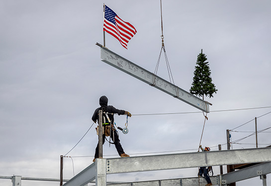 A worker stands atop the steel frame of a partly constructed building, guiding into place a large steel beam being held by a crane, an American flag