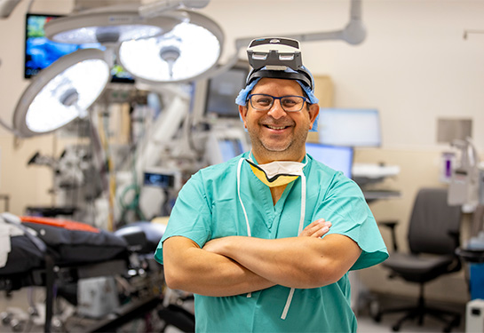 A surgeon wearing green scrubs, a cap and an augmented reality headset stands in an operating room.