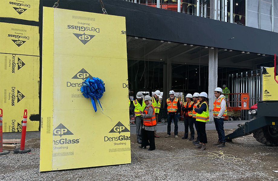 A group of people in construction hats watch a large yellow wall pane lifted for installation on a building under construction
