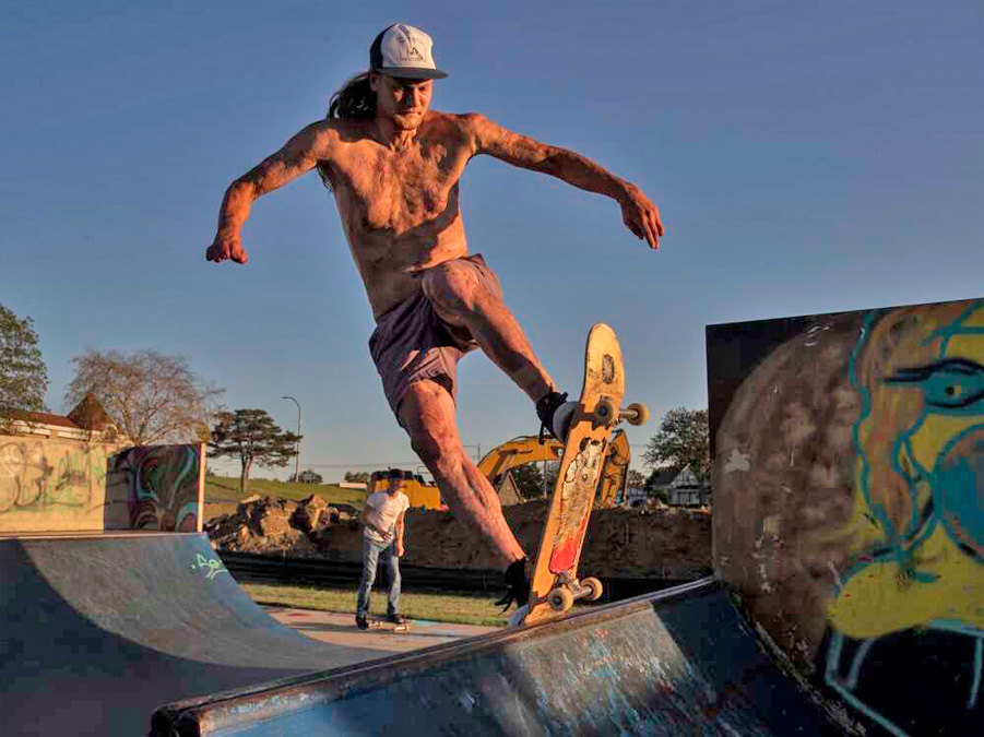 A shirtless man wearing baseball cap, shorts and showing visible burn scars rides a skateboard up a ramp at a skateboard park