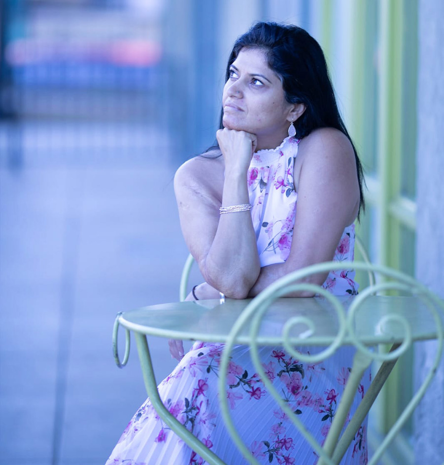 Woman in floral dress sits at an outdoor metal table leaning on her right elbow and looking toward the sky.