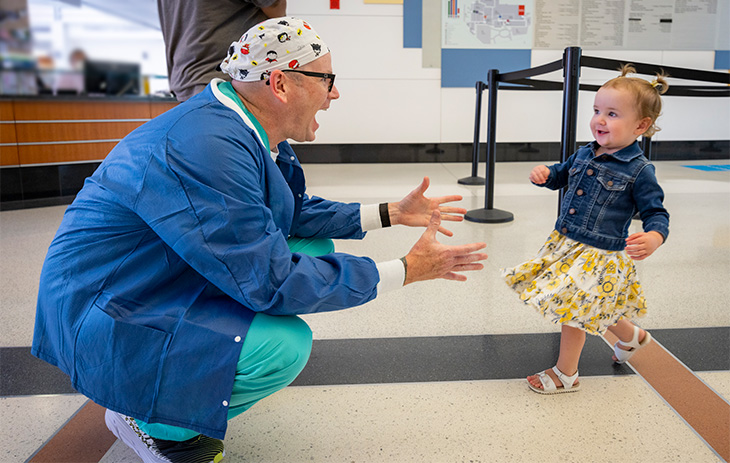 Man in nurse uniform holds arms out and toddler girl with pigtails runs toward him smiling.