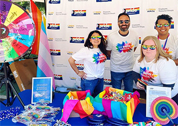 An outdoor stand with four persons wearing white T-shirts with a blast of colors in the center and UC Davis Health writing. Behind them is a white board with UC Davis Health logo, Employee Resource Groups and the pride flag. 