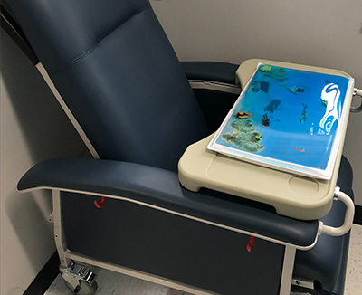 A large blue, padded medical chair with wheels and a blue tray featuring sea creatures sits in the corner of a room.