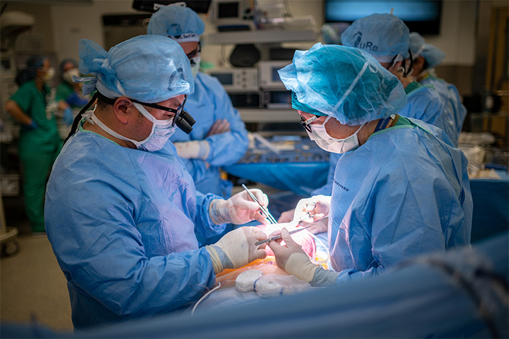 Two surgeons in blue scrubs, masks and head covers are operating on a patient, with other medical team members observing in the operating room.