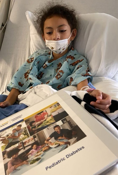Elena in a hospital bed with a book 'pediatric diabetes' on her lap
