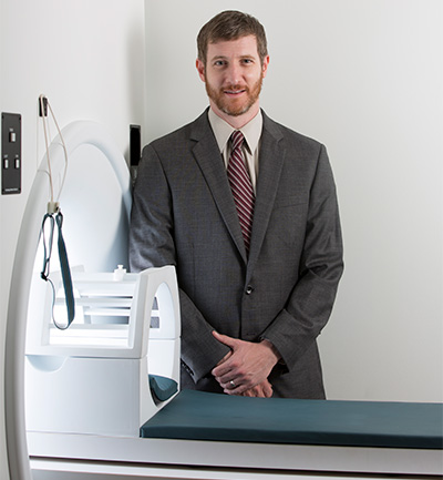 A brown-haired man in a gray suit with cream-colored shirt and burgundy tie stands next to an MRI machine. 