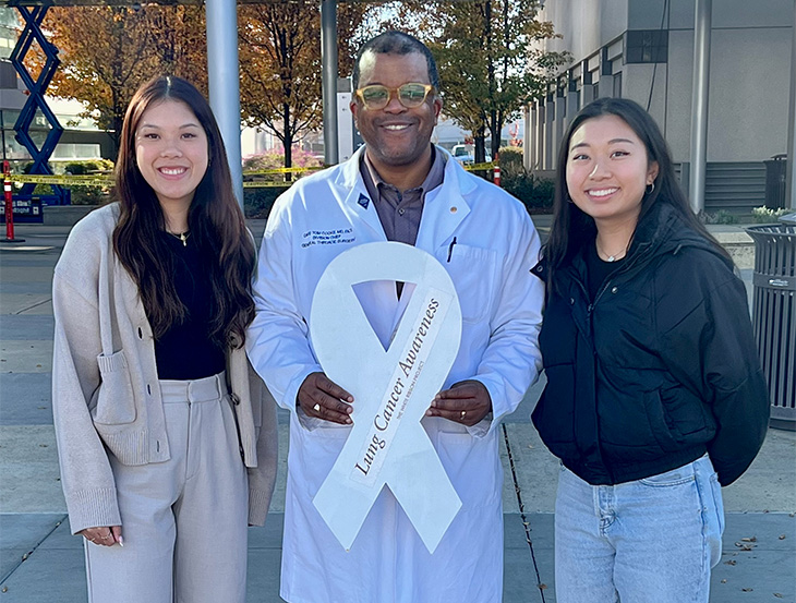 Man in a white doctor’s coat accepts two-foot-tall white ribbon from two female students standing next to him. 