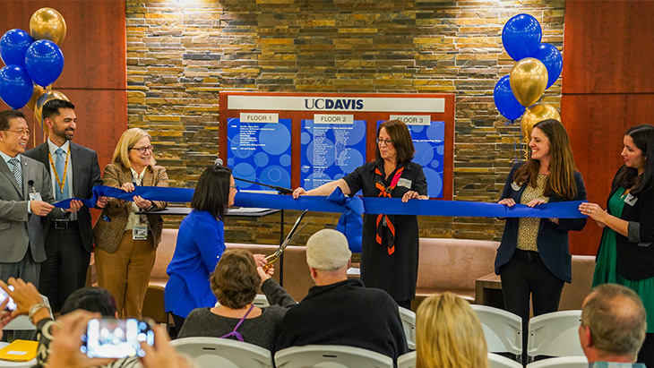 Woman in a blue blouse uses giant gold scissors to cut a blue ceremonial ribbon held by colleagues.