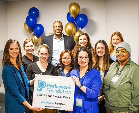 A diverse group of people stands together holding a “Parkinson’s Foundation Center of Excellence” sign amid blue and gold balloons.