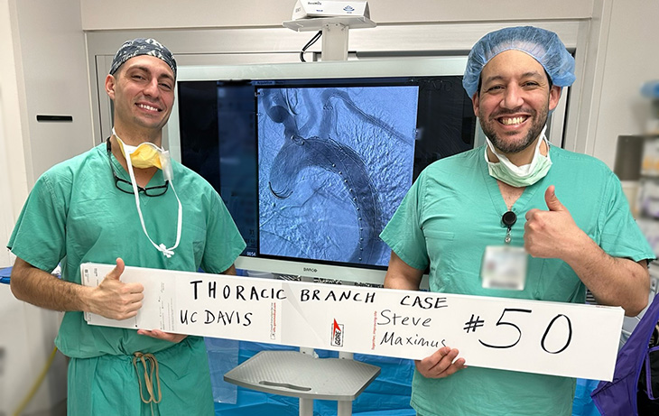 Two doctors in green scrubs stand in front of a screen showing a medical scan, holding a white sign that says “Thoracic Branch case #50, UC Davis.”