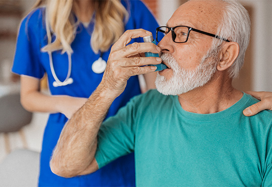 Man with white beard and green shirt breathing in medication with an inhaler as a nurse with blue scrubs standing behind him.