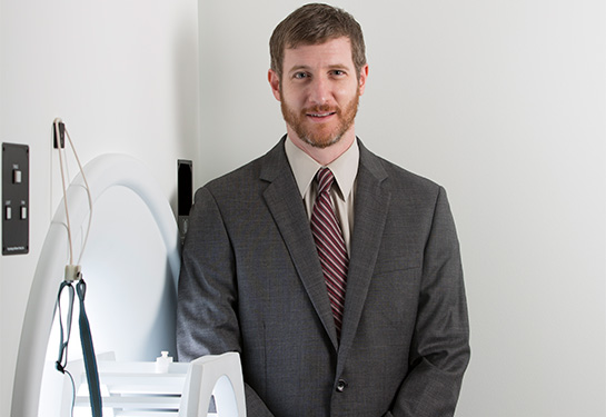 A brown-haired man in a gray suit with cream-colored shirt and burgundy tie stands next to an MRI machine. 