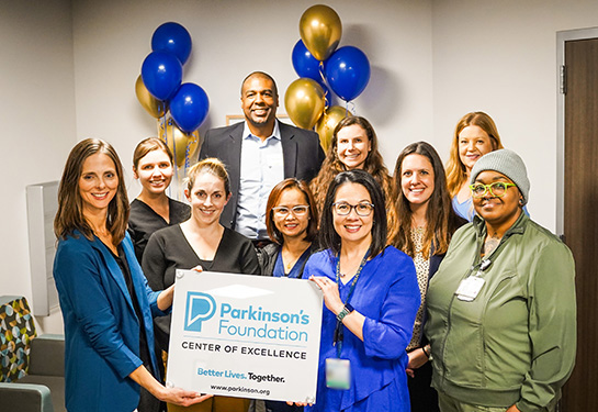 A diverse group of people stands together holding a &#x201c;Parkinson&#x2019;s Foundation Center of Excellence&#x201d; sign amid blue and gold balloons.