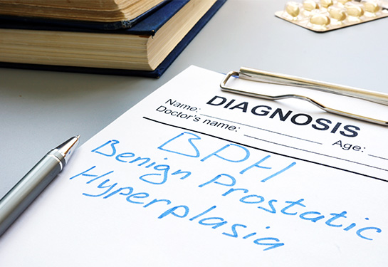 A clipboard holding white paper and the terms &#x201c;diagnosis, Benign prostatic hyperplasia&#x201d; with a silver pen, packet of yellow pills and two large books