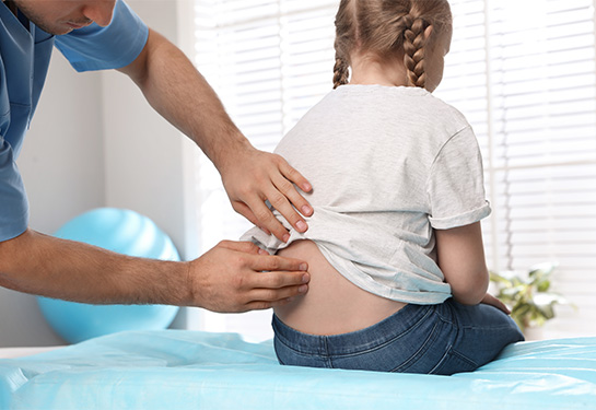 A person wearing blue scrubs and examining the back of a child. The child has blonde braids, wearing white shirt and blue jeans