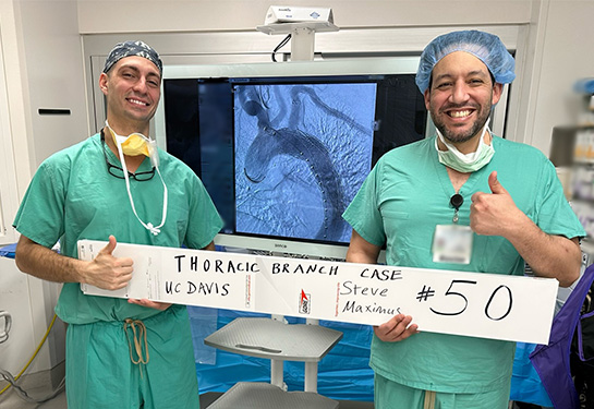 Two doctors in green scrubs stand in front of a screen showing a medical scan, holding a white sign that says &#x201c;Thoracic Branch case #50, UC Davis.&#x201d;