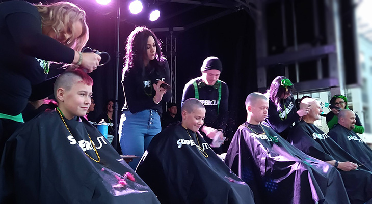 Three young boys and two men sitting on stage, wearing dark drapes, getting their hair shaved by four people.