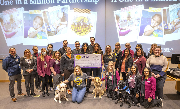 Twenty-four people and three dogs gather in a room holding an oversized check with $90,600 written on it