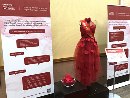A long red dress with red hearts is displayed on a mannequin at a table between two tall vinyl posters.
