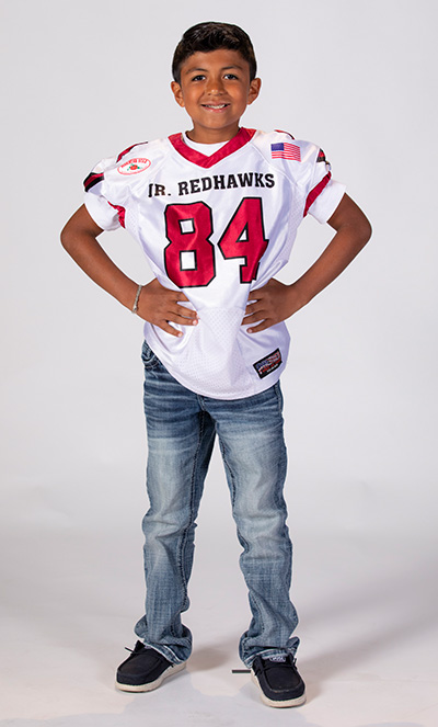 Boy in blue jeans and white football jersey with the number 84 in red, standing with his hands on his hips.