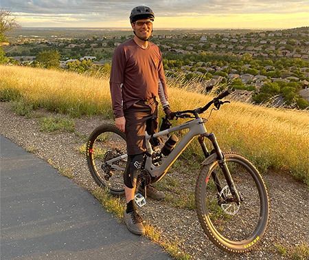 Man with brown hair standing next to bicycle parked on trail surrounded by grass and a residential development in the background.