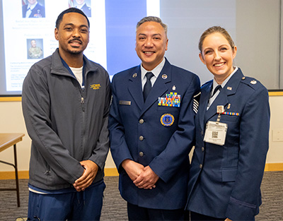 Three people, one in blue pants and a gray jacket, two in Air Force uniforms, stand together for a photo.