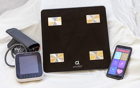 Black scale, black handheld tablet and gray blood pressure cuff on white sheet.