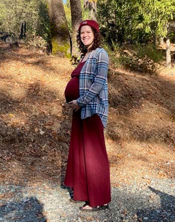 A pregnant woman in a long maroon dress stands outside with tall trees in the background