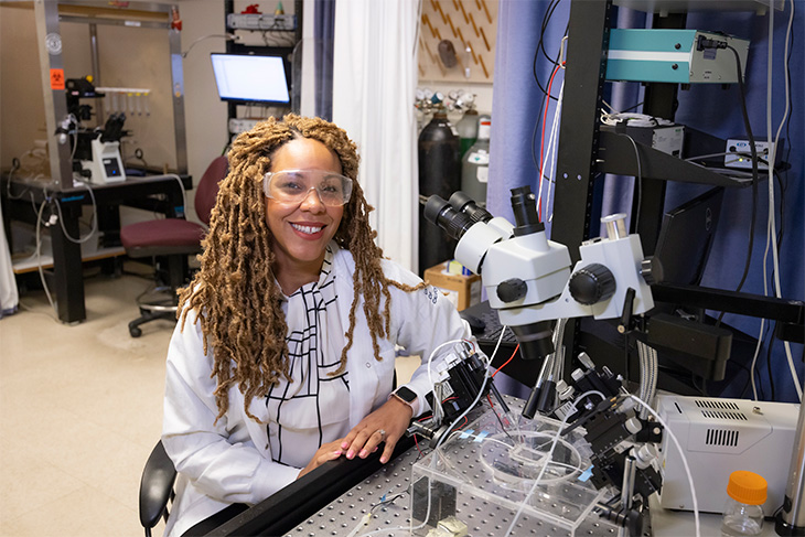 A smiling woman with long brown hair sits in front of a microscope wearing goggles and a white lab coat.