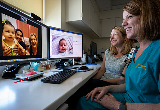 A woman in teal scrubs seated next to a woman in a green shirt watch a computer screen that shows a man and woman holding a baby 