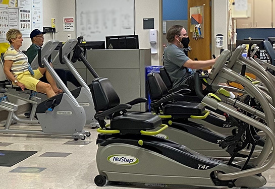 Three people sitting on exercise bikes in exercise room with three people in medical scrubs standing in the back of the room.