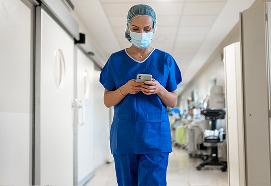 A masked female physician wearing blue scrubs looks down at a smartphone while walking down a hospital corridor.