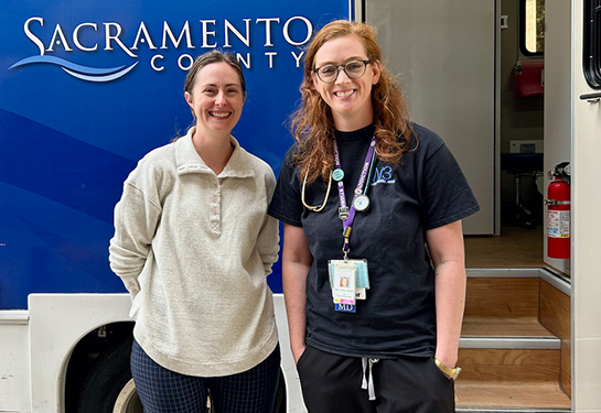 A woman in a beige sweater stands next to a woman in a blue shirt outside an RV with the words “Sacramento County” on the side