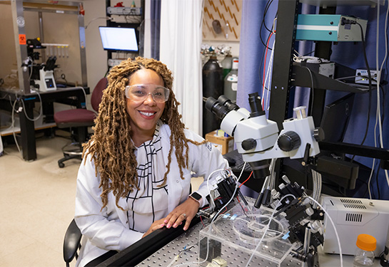 A smiling woman with long brown hair sits in front of a microscope wearing goggles and a white lab coat. 