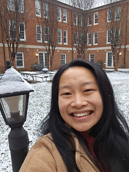 A woman with dark hair past her shoulders smiles in front of a red brick building in a courtyard where snow covers the ground